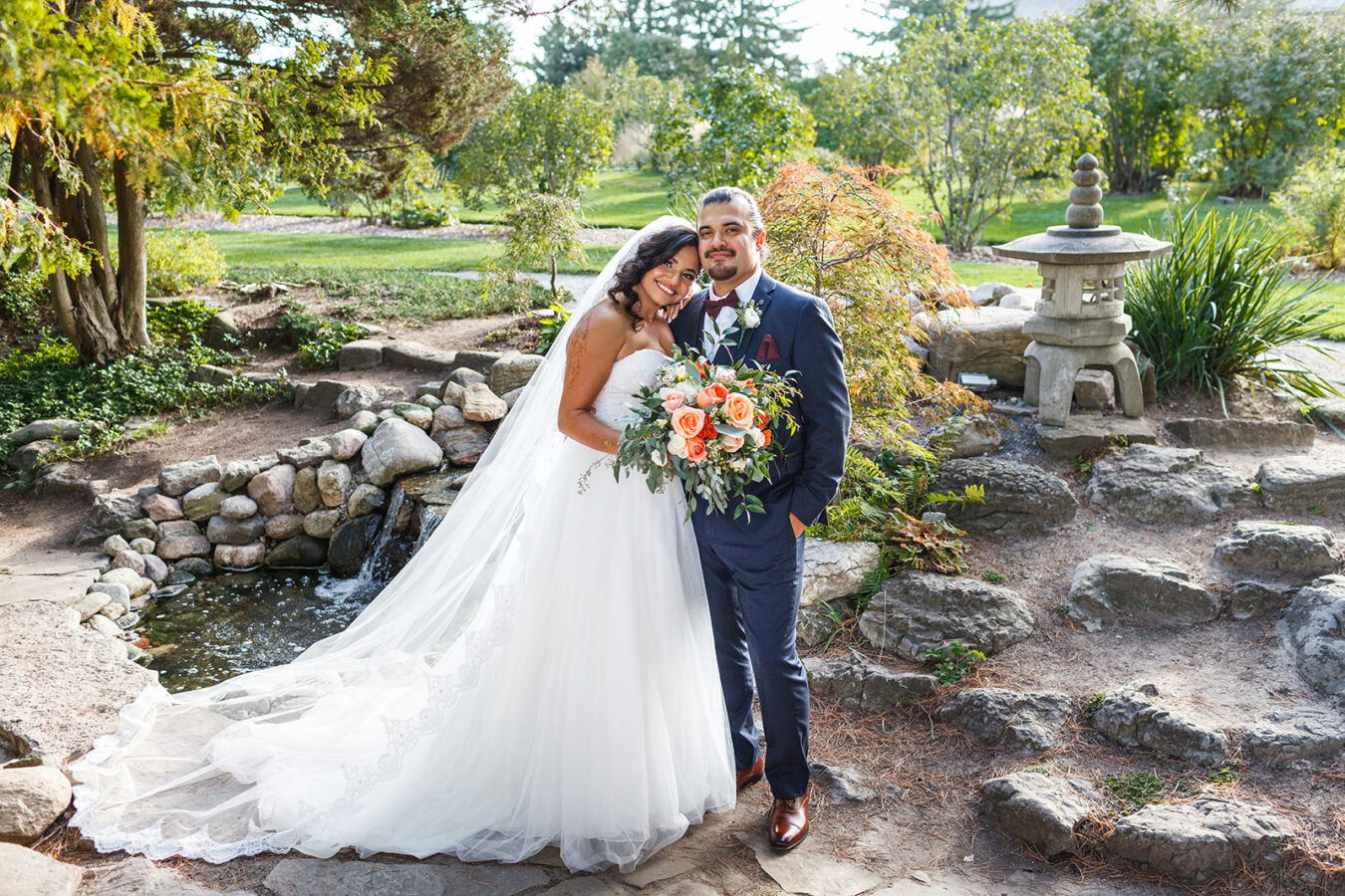 A couple stands together on their Ottawa wedding day. The bride wears a white dress and the foliage is green with a river and garden in the background.
