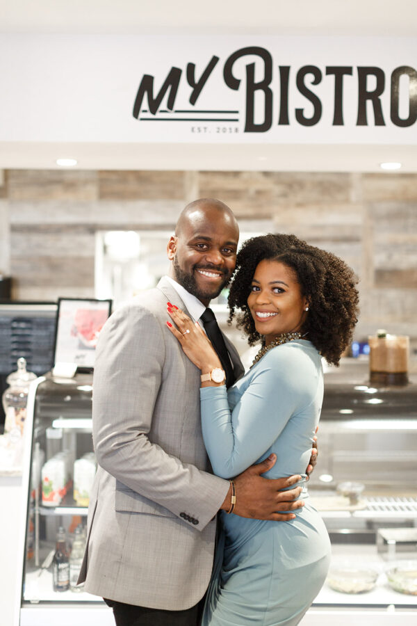 A couple stand in a coffee shop under a sign that says my bistro smiling for their engagement pictures