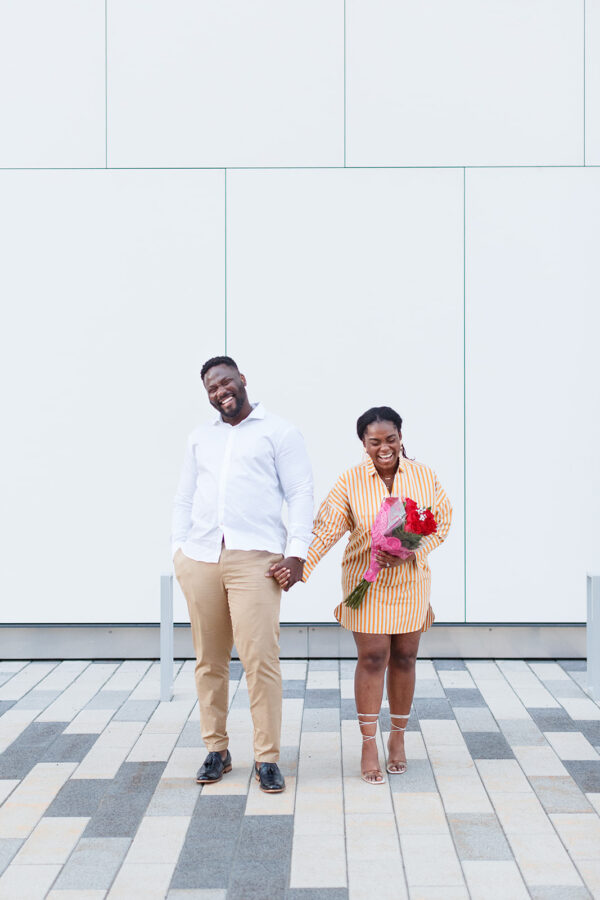 A couple stand in front of a white wall in peach and white she is holding a fuschia flower boquet and they are laughing