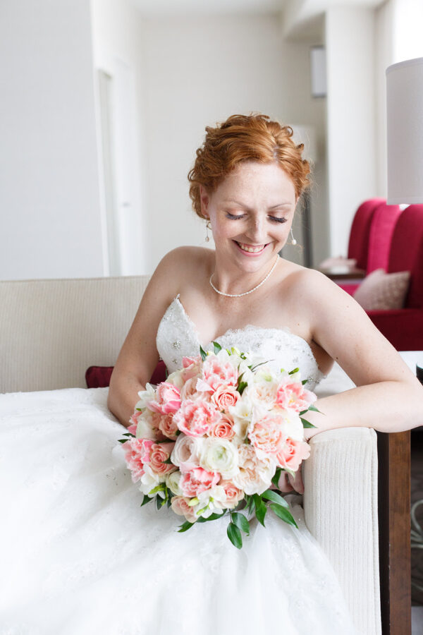 A bride with red hair smiles and looks down at her pale pink boquet